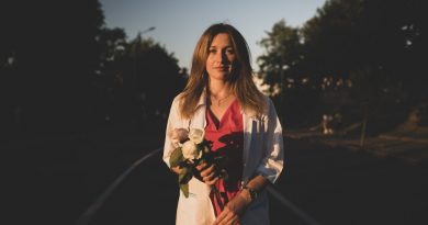 Photo depicts a woman dressed in a white jacket and a red shirt, holding white roses.