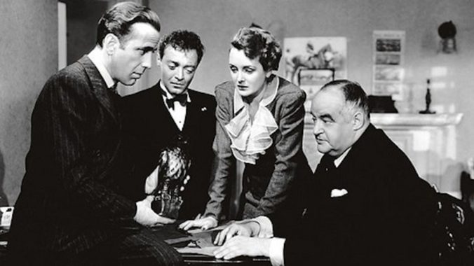 In a black and white move, four actors stand around a table in deep conversation. In one actor's hands is a small black falcon statue. The three other actors are looking at the statue.