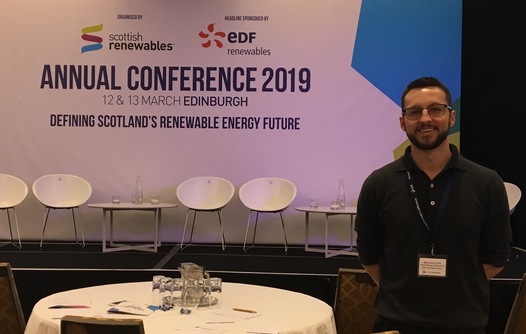 Iain standing to the right of a round table, with a stage in the background that has white chairs and a backdrop reading "Annual Conference 2019"