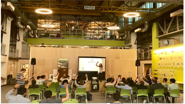 Photo of a presentation in an industrial space, attendees are sitting in green chairs and raising their hands as someone stands in front of a screen speaking into a microphone