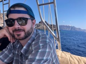 Photo of Matt Arnold wearing a backwards cap and sunglasses, with ocean in the background