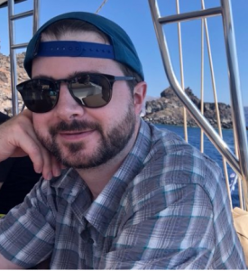 Photo of Matt Arnold wearing a backwards cap and sunglasses, with ocean in the background
