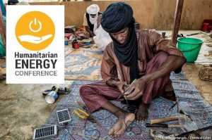 Man wearing a turban scarf sitting on a rug working on electronics, Humanitarian Energy Conference logo in the upper left hand corner