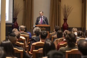 Photo of Adnan Amin standing at a podium in front of a room of seated people, speaking into a microphone