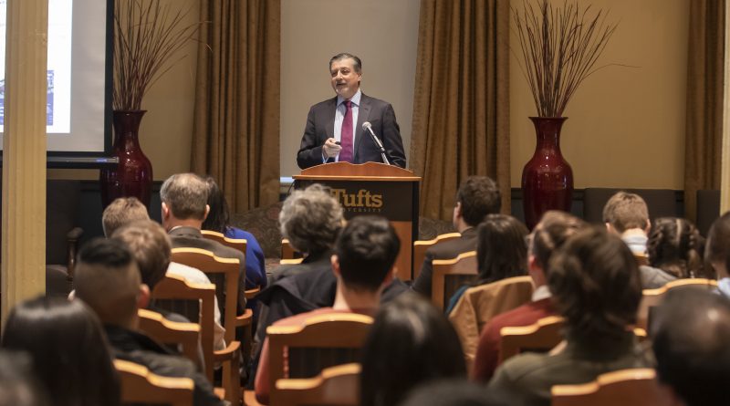 Photo of Adnan Amin standing at a podium in front of a room of seated people, speaking into a microphone