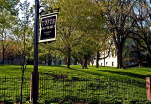 Photo of a street sign for Tufts University with trees, a hill, and a campus building in the background