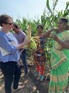 Kelly Sims Gallagher and Rishi Bhandary stand in a cornfield with two women in India. Kelly is holding a shucked ear of corn and speaking to one of the women.