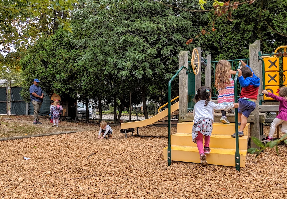 a group of children climbing up steps of playground structure, one child touching the mulch ground cover, and in background teacher talking to a group of children