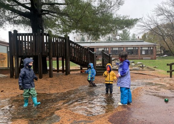 Four children in raingear playing in rain puddle next to a wooden tree house on a cloudy day