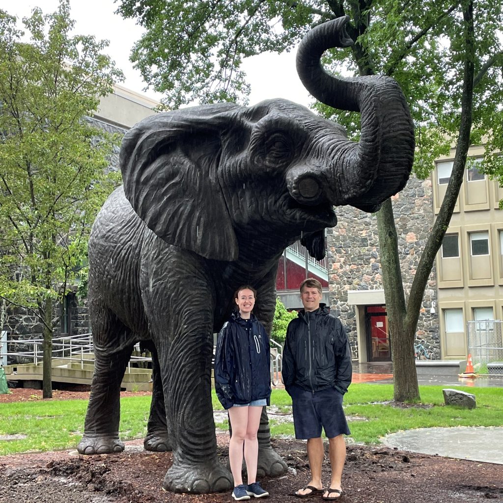 Colleen J. and Mat G. pose in front of the Tufts Jumbo mascot on the Tufts campus in the rain. 