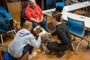 an older person sitting in a chair while two young individuals pet her therapy dog.