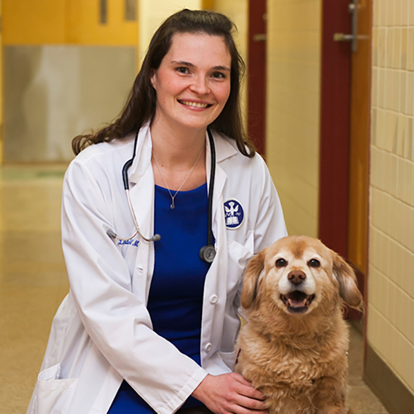 a smiling individual with long dark hair wearing a white medical coat kneeling beside a medium sized beige dog