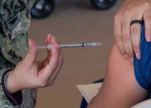 Close up of a person's hand administering a vaccination into the arm of another person. The person giving the vaccine is wearing military fatigues