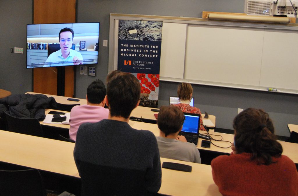 Photo of Deltan in a white shirt on a large tv screen speaking to students who are sitting in a classroom
