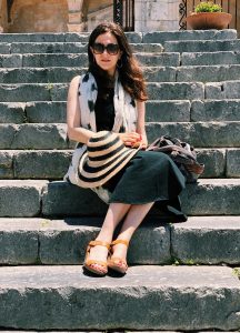 Jennie Hirsh, a woman with long brown hair, sits on stairs. She wears a black and white outfit with yellow shoes.