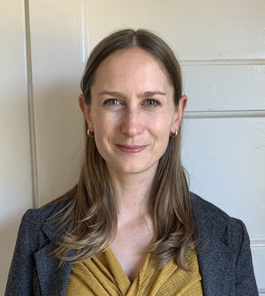 Photo of a person with long brown hair with a middle part. They wear a gray blazer and yellow shirt. 