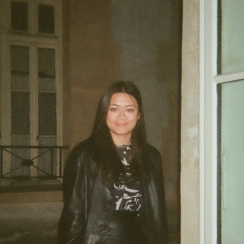 CoCo looks at the camera and smiles. She wears all black and stands in front of a brick building with thin rectangular windows.