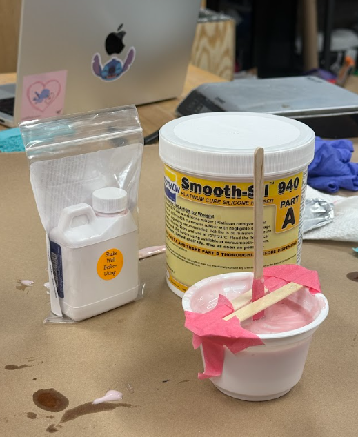 A sacrificial mold made with a yogurt cup. The cup contains unsolidified Smooth-Sil 940. Two popsicle sticks are holding a longer popsicle stick that has the 3D printed wedge submerged.