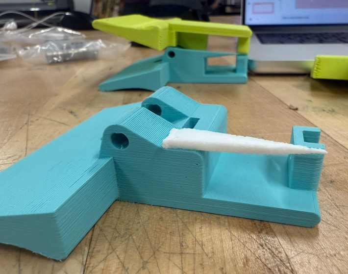 One half of our device, shown in teal, laid out on a table. The wedge of opening mechanism is made with white polyurethane. The photo is taken from a side-view, highlighting the thickness and pointiness of our opening mechanism.