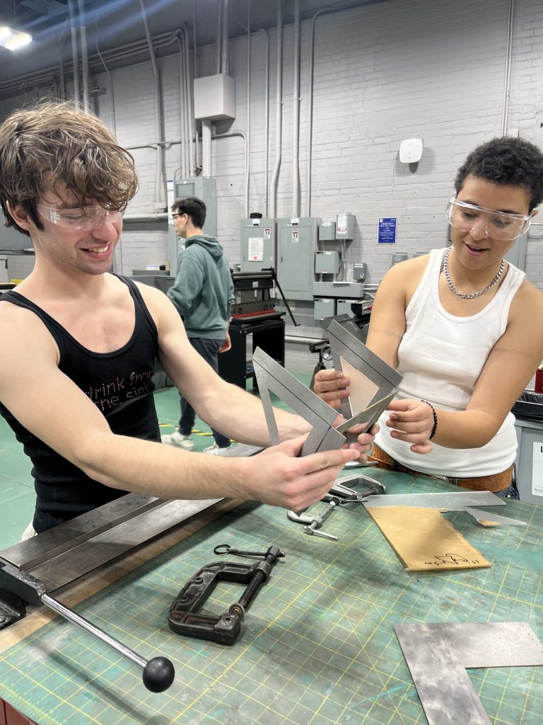 Two team members in the Bray Lab working on manufacturing a lamp shelf. They are both holding the skeleton of the shelf and examining it, surrounded by materials and equipment in a machine shop