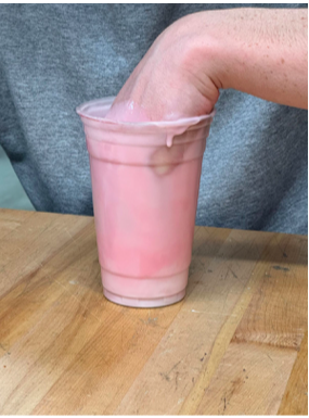 Picture including a cup on a wood table with a hand inside the mixed pink molding material.