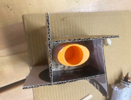 Picture including an orange elliptical 3D printed object with a hole inside a cardboard box.