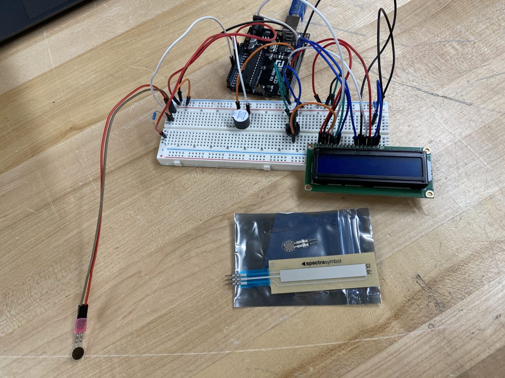 Bread board with pressure sensor, buzzer, LCD, and arduino connected by wiring with additional sensors on the table.