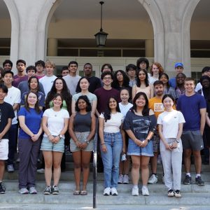 Image of a group of about 60 students standing in rows on the steps of a pale-colored building in the background