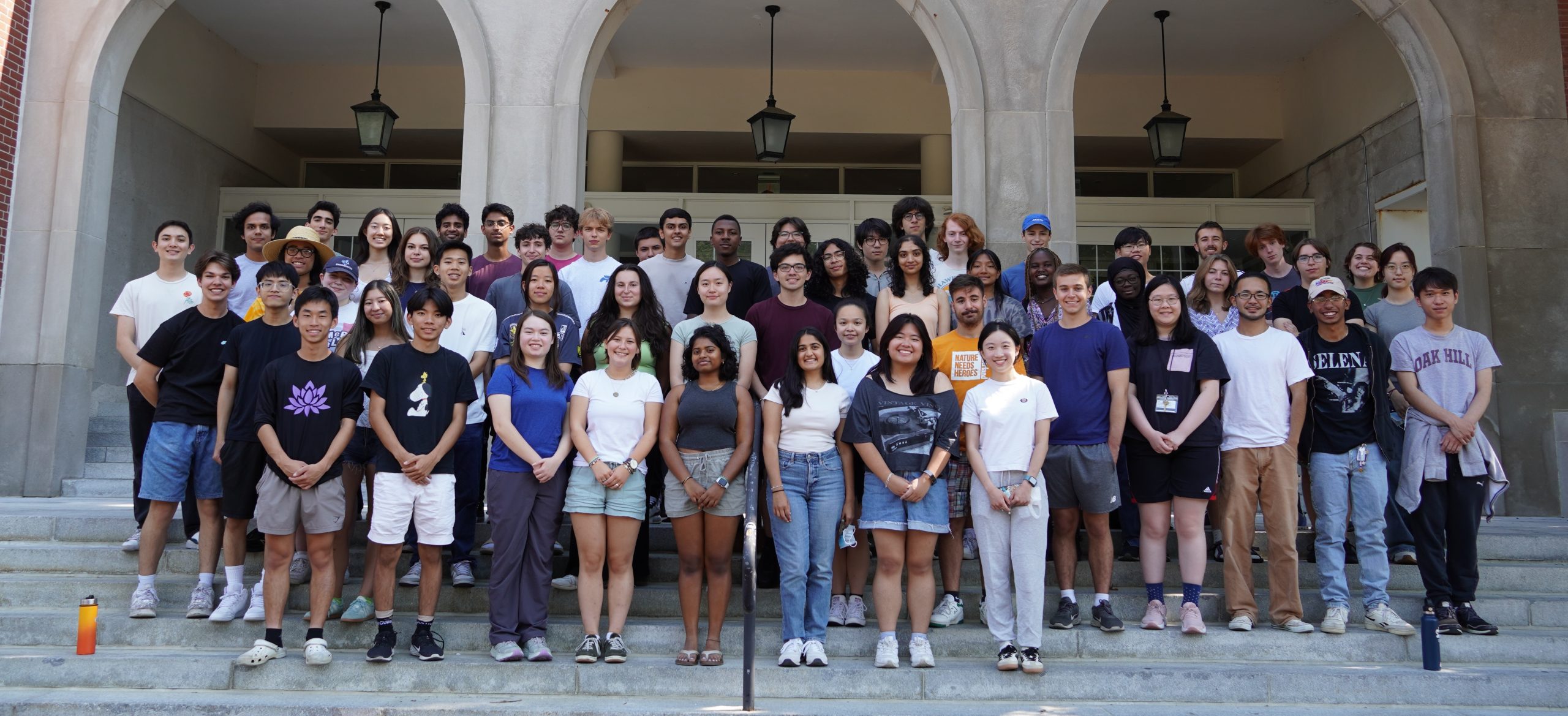 Image of a group of about 60 students standing in rows on the steps of a pale-colored building in the background