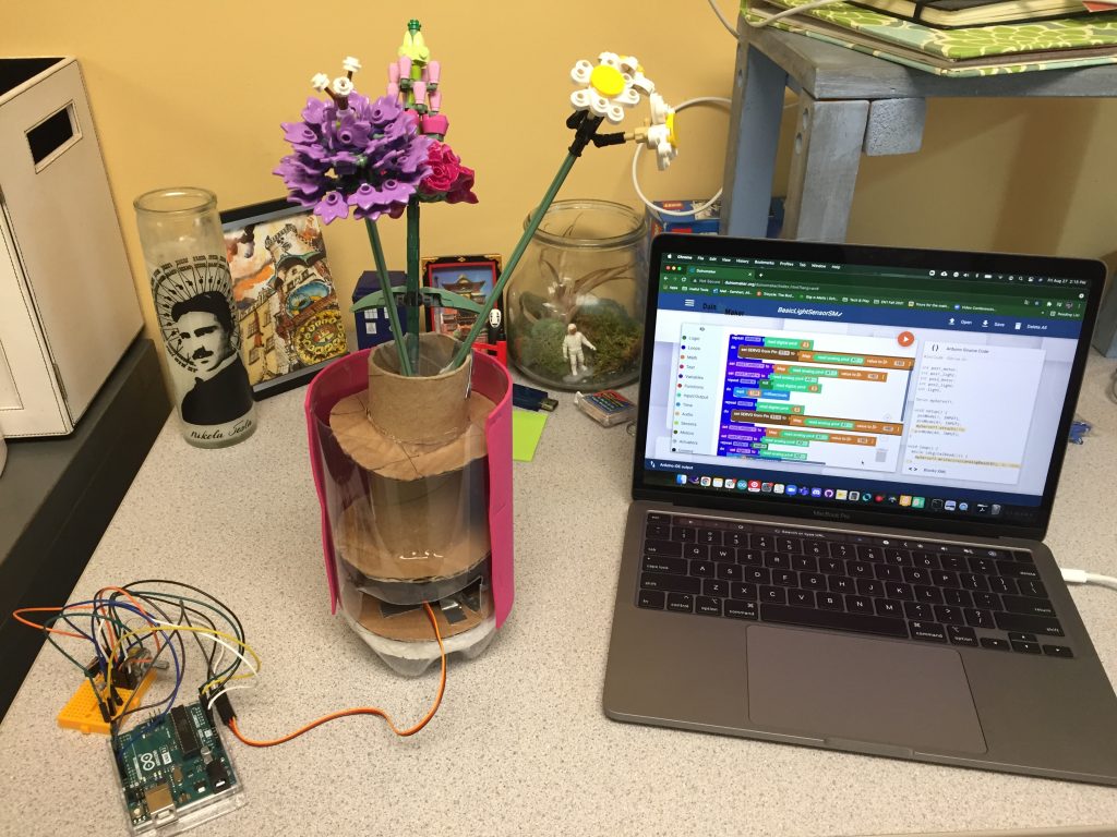 Three LEGO flowers in an improvised vase made of a plastic bottle and some cardboard. The vase sits on a desk next to a computer showing the Duinomaker block code, and an Arduino with some electronics connected to it. 