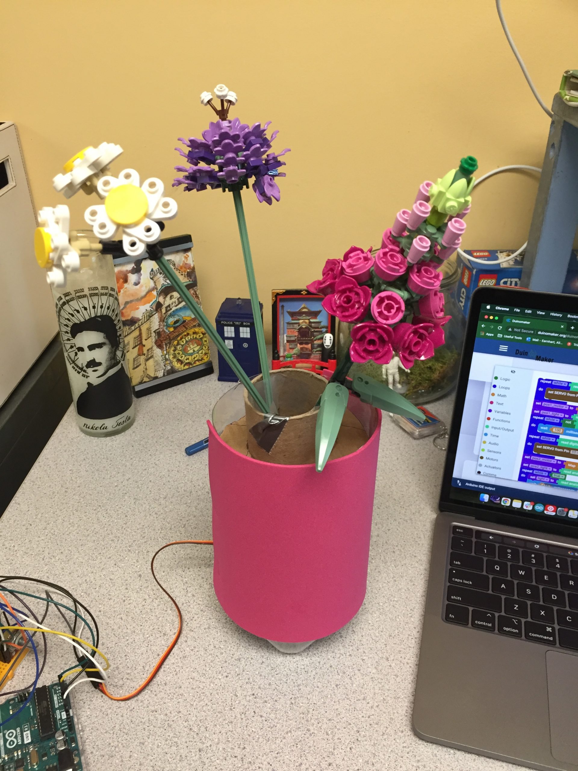 Flowers made of LEGO in a vase on a desk, next to a computer and some electronics components.