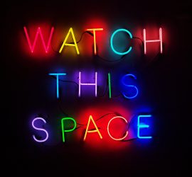 A multi-colored neon sign, on a black background, that says "watch this space."