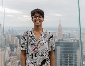 A smiling girl with dark brown hair and brown skin and glasses, with a city skyline in the background