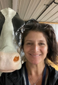 A light-skinned woman with brown hair smiling, with a simulated cow head over her shoulder.