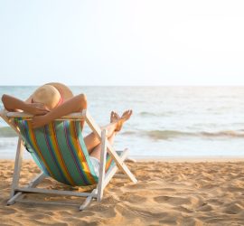 A woman with hat relaxing and arm up on beach chair on the beach, looking at the ocean in front of her.