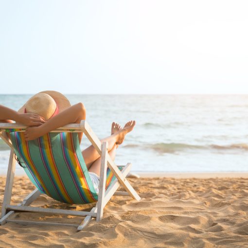 A woman with hat relaxing and arm up on beach chair on the beach, looking at the ocean in front of her.