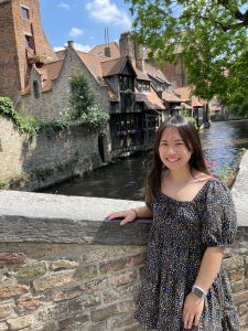 A young woman smiles while standing by a stone wall with historic, canal-side buildings in the background on a sunny day.