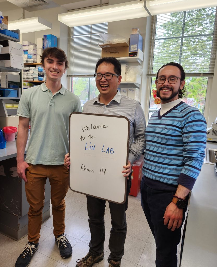 Yi-Pin, Connor and Sergio stand inside the lab with a sign that reads "Welcome to the Lin Lab, Room 117"