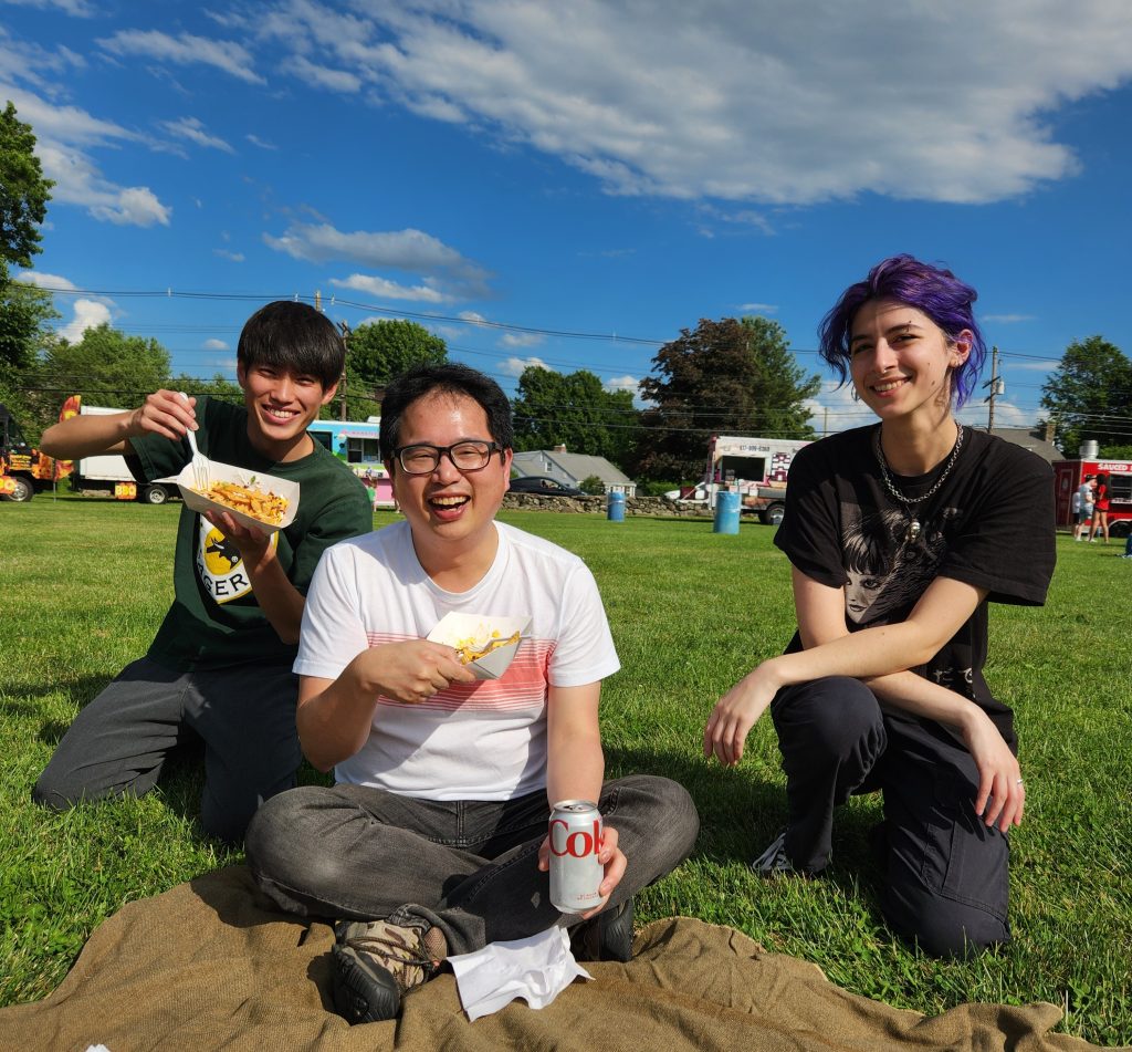 Yi-Pin, Takeuchi (PhD student Summer 2024), and Carly sit outside on grass on a sunny day. Yi-Pin is holding fries and a  can of coke, and Take is holding fries and a fork.