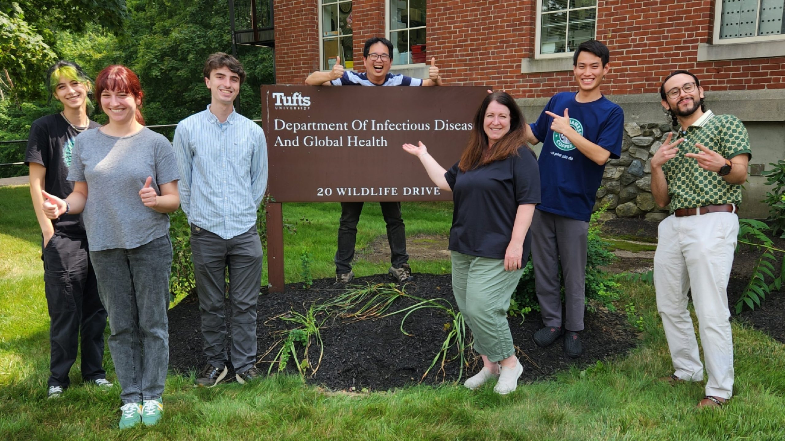 The Lin Lab stands outside in front of a sign that reads, "Tufts University: Department of Infectious Disease and Global Health; 20 Wildlife Drive." From left to right: Carly, Miranda, Connor, Yi-Pin, Loranne, Take, Sergio.